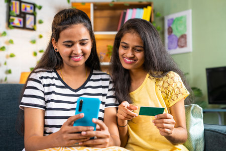 Happy smiling young siblings making online payment for shopping on mobile phone by using credit card at home - concept of e commerce, online banking and bondingの写真素材