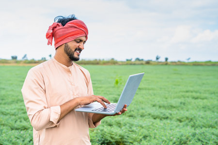 Young indian farmer busy wrking on laptop while sitting at agricultural farmland - concept of technology, digital communication and modern farming.の写真素材