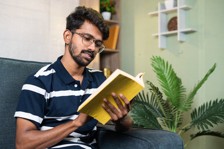 serious young indian student reading book during free time at home - concept of education, hobbies and hard worker.の写真素材
