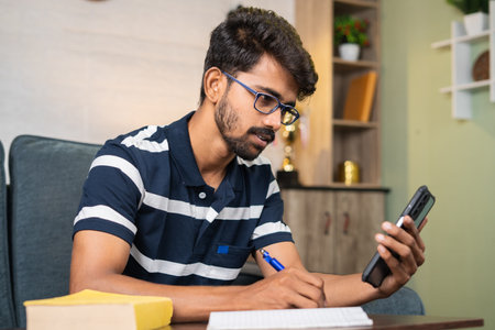 Young man using mobile phone for writing notes at home while listening online class - concept of...