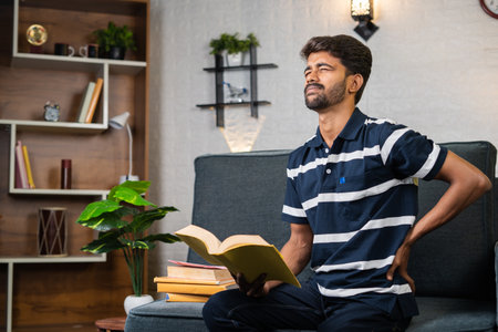 young student at home suffering from back pain while reading book due long time reading on sofa -...