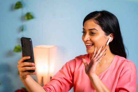 Happy young girl with earbuds talking in video call on mobile phone at home - concept of technology, connection and communication.の写真素材