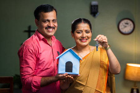 Happy smiling couple showing new house keys by toy home while looking camera - concept of new home purchasing, home loan and investment.の写真素材