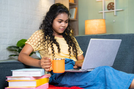 Young indian girl drinking coffee or tea while using laptop during examination while sitting on sofa at home with books - concept of refreshment, freelancer and online shoppingの写真素材