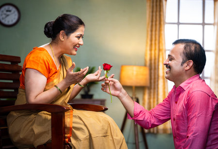 Happy middle aged man proposing his wife by giving red rose at home - concept of wedding...