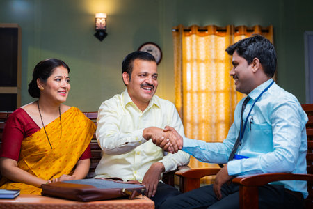 Banking officer greeting by shaking hand to couple by explaining about insurance at home - concept...