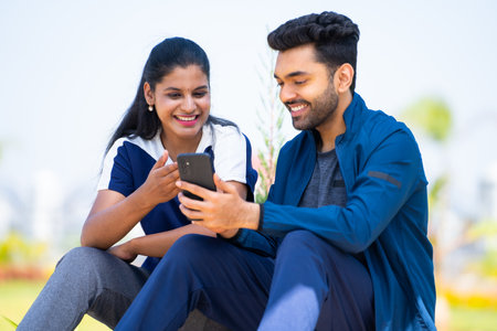 Happy young man showing mobile phone to girlfriend while sitting at park during morning walk - concept of social media sharing, active wear and using fitness app.の写真素材