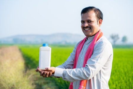 Happy Indian village farmer holding fertilizer bottle by looking at camera at farmland - concept of product promotion, recommendation and agriculturalの写真素材