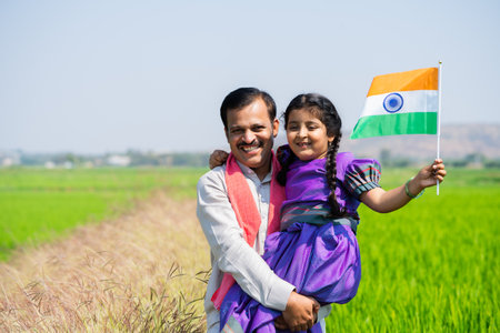 Happy Indian village father carrying daughter with indian flag by walking near green farmland during independence day - concept of democracy, patriotism and relationship bonding.の写真素材