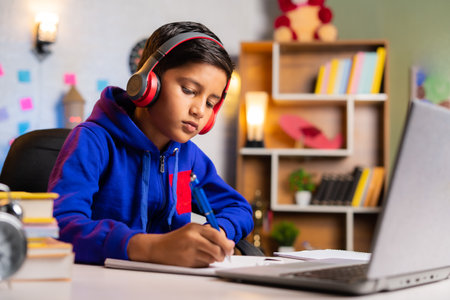 Indian kid making notes from online class on green screen laptop at home - concept of e-learning,...