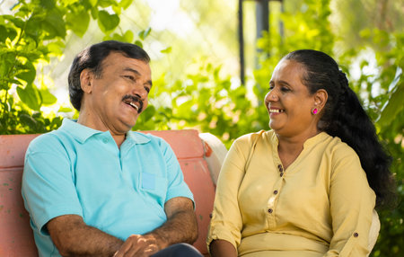 Happy indian senior couple talking each other while sitting at park - concept of relaxation,...