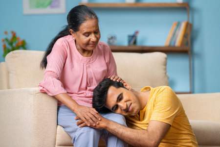 Indian senior mothers talking to adult son by consoling while son sleeping on mothers lap at home - concept of motherhood, parental support and gratitudeの写真素材