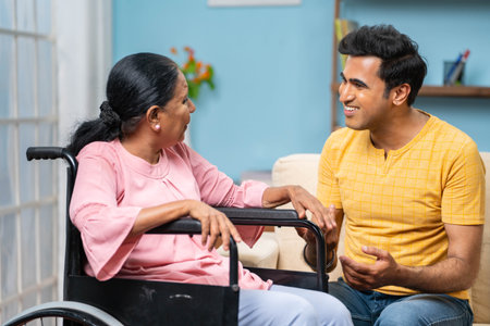 Happy indian adult son with disabled senior mother on wheelchair spending time by talking at home -...