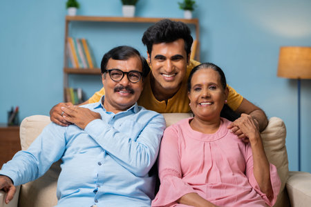 portrait shot of Happy smiling indian senior parents with adult son looking at camera while sitting on sofa at home - concept of family bonding, relationship and relaxation.の写真素材