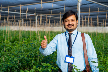 Happy smiling indian banker or officer standing by showing thumbs up while looking camera at greenhouse - concept of successful, banking support and environmentalの写真素材