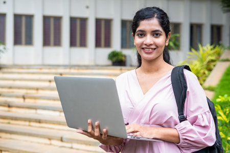 Confident girl student with backpack holding laptop by looking at camera at college campus - concept of education, technology and developmentの写真素材