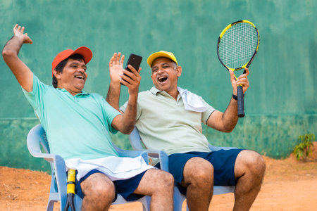 Happy laughing indian senior friends at tennis court by watching mobile phone - concept of taking break, social media and technologyの写真素材