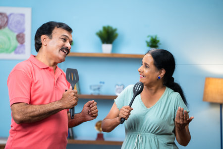 Happy indian senior couple dancing and singing by holding cooking spoons at home - concept of active retirement lifestyles, love emotion and healthy relationshipの写真素材