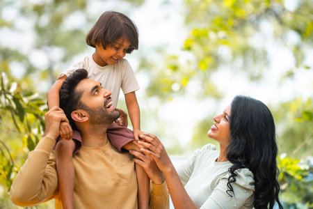 Happy indian parents playing with daughter while kid on father shoulder at outdoor - concept of parenthood, family bonding and weekend holidaysの写真素材