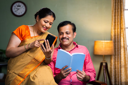 Happy indian woman showing mobile phone to husband while reading book at home - concept of technology, togetherness and social media.の写真素材