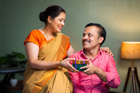 Happy indian woman giving gift to husband at home - concept of wedding anniversary, togetherness and affectionの写真素材