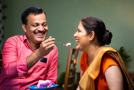 Happy Indian husband feeding cake to wife while sitting on sofa at home - concept of wedding anniversary celebrating, family bonding and affection.の写真素材