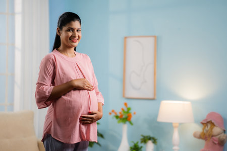 Happy smiling Indian Pregnant woman standing by feeling tummy while looking camera at home - concept of motherhood, expecting baby and parenthoodの写真素材