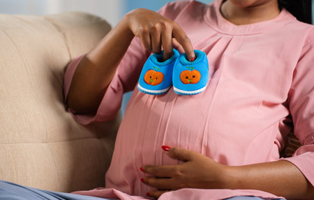 Close up headless shot of Indian pregnant woman playin by placing baby booties on tummy at home - concept of orenatal connection, motherhood and pregnancyの写真素材