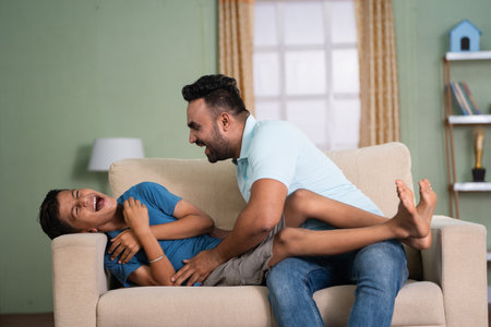 joyful indian father playing by cuddling his son on sofa at home - concept of family bonding, spending time and affectionate.の写真素材
