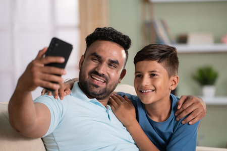 Happy smiling indian father with son taking selfie on mobile phone on sofa at home - concept of social media sharing, cyberspace and family time.の写真素材