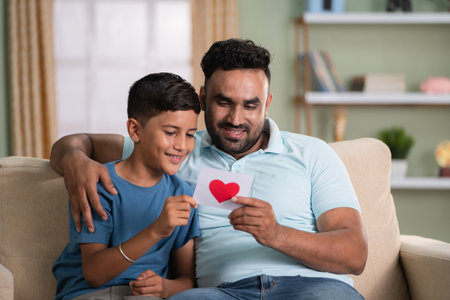 Happy smiling indian son giving greeting card to father by embracing while sitting on sofa at home - concept of family relationship, affectionate and fathers dayの写真素材