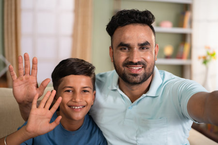 Happy smiling indian father with son talking video call by holding camera at home - concept of long distance relationship, social media live and cyberspaceの写真素材