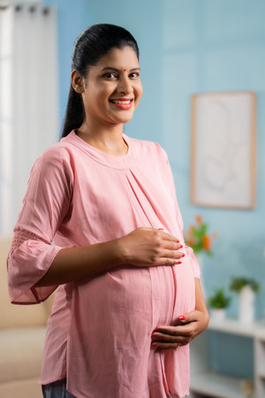 vertical Portrait shot of happy smiling Indian Pregnant woman standing by holding tummy by looking at camera - concept of motherhood, expecting baby and parenthoodの写真素材