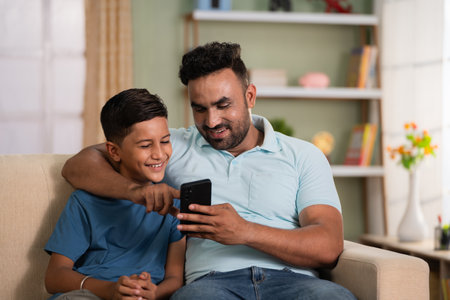 Happy smiling indian father showing mobile phone to son while sitting on sofa at home - concept of technology, togetherness and fatherhood.の写真素材