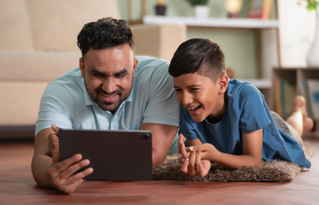 joyful indian father with son using digital tablet while lying on floor at home - concept of weekend holiday, Modern parenting and Digital entertainmentの写真素材