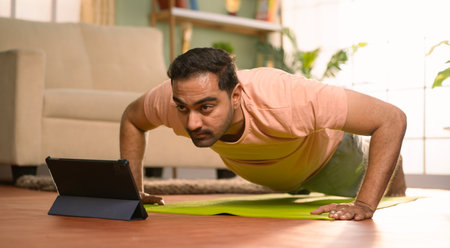 Indian man doing push ups exercise by online video classes from tablet at home - concept of Fitness instruction, Exercise Tutorial and Bodyweight Trainingの写真素材