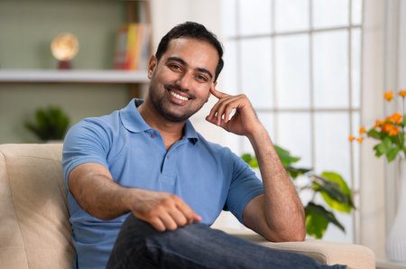 Happy smiling relaxed indian man looking camera while sitting on sofa at home - concept of Comfortable leisure, satisfied lifestyle and carefree living.の写真素材