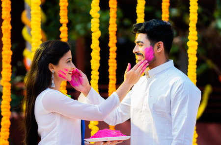 Happy indian young couple applying holi colors on face together by holding holi power plate flower decoration background - concept of holi festival celebration, relationship and Indian tradition culture.の写真素材
