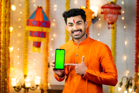 Happy smiling indian man in traditional ethnic wear showing green screen mobile phone by pointing finger by looking camera - concept of app or application, promotion, festival sales and offersの写真素材