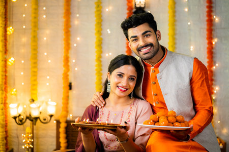 Happy smiling indian couple in traditional ethnic wear by holding sweets at home - concept of diwali festive celebration, greeting and family relationshipの写真素材