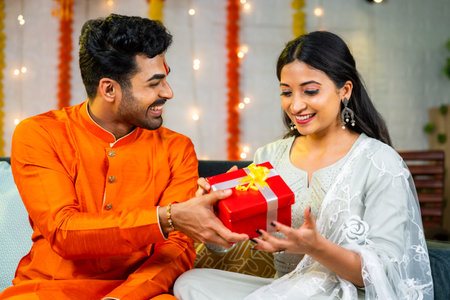 Happy young husband giving present to wife during festival celebration at home - brother giving gift to sister during raksha bandhan.の写真素材