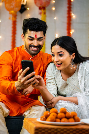 Vertical shot of happy indian couple using mobile phone during festival celebration at home - concept of festival sale, discounts or offers and social media sharingの写真素材