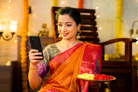 Medium shot of happy indian girl using mobile phone to check for new decorative designs by holding flowers plate - concept of technology, festive planning and social media sharing.の写真素材