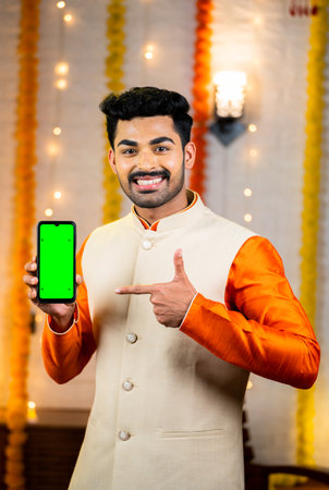 vertical shot happy indian man in traditional ethnic wear showing green screen mobile phone by pointing finger by looking camera - concept of app or application, promotion, festival sales and offersの写真素材