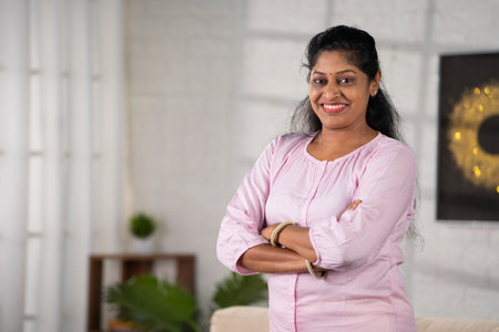 Happy smiling middle aged indian woman standing with arms crossed by looking camera at home with copy space - concept of successful, empowerment and inspirational.の写真素材