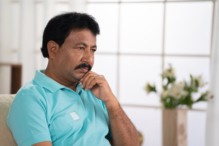 Thoughtful worried indian middled aged man deep thinking while sitting on sofa at home - concept of loneliness, financial problems and mental illnessの写真素材