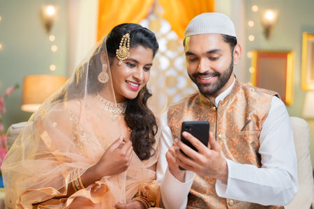 Happy smiling indian Muslim couple busy using mobile phone during Ramadan festival sales at home - concept of social media sharing, technology and family bonding.の写真素材