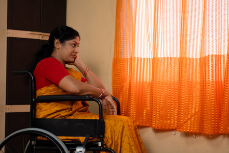 Alone Indian thoughtful senior woman sitting on wheelchair by looking window at home - concept of rehabilitation, trauma and mental well beingの写真素材