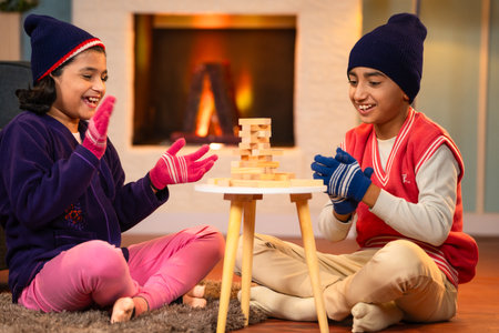 Indian siblings kids in winter wear playing wooden blocks building game at home during winter holidays at home - concept of childhood concentration, entertainment and relationship bondingの写真素材