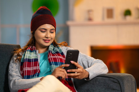 Happy young woman in winter wear busy using mobile phone at home - concept of social media sharing,...